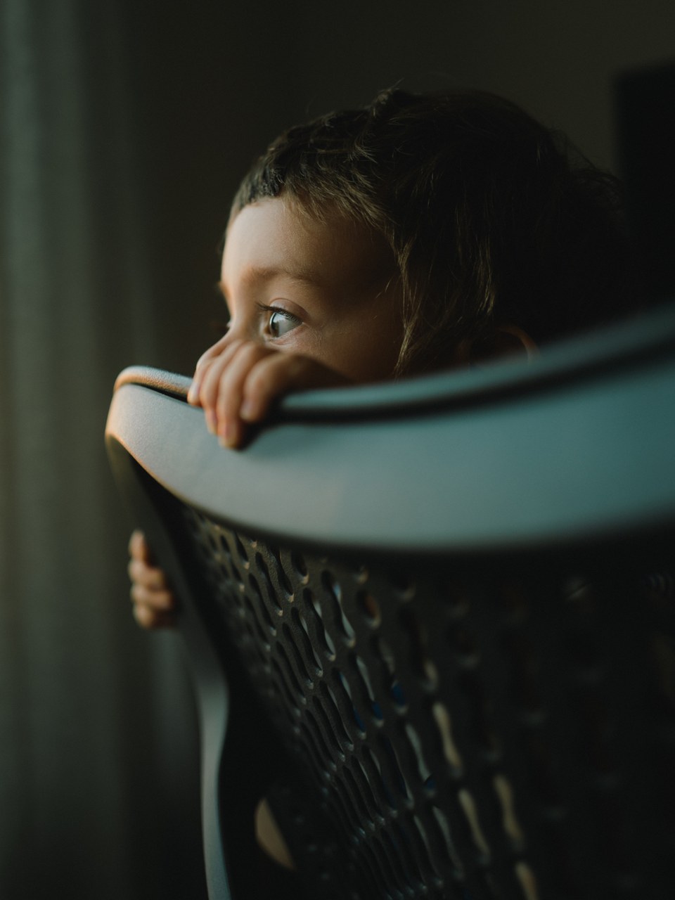 Toddler peeking over the edge of a chair in dim window light